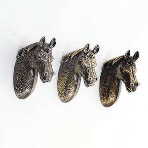 Puede incluir: Tres ganchos de pared con forma de cabeza de caballo en diferentes acabados: negro, bronce y dorado. Los ganchos están hechos de metal y tienen un diseño realista de cabeza de caballo.