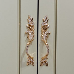 Może przedstawiać: Dwa ozdobne złote i srebrne uchwyty do drzwi szafki z kwiatowym wzorem.