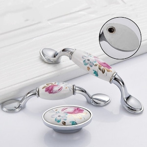 Op de afbeelding: Twee witte keramische kastknoppen met bloemmotieven en zilveren accenten. De knoppen zijn ovaalvormig en hebben een gebogen handvat.