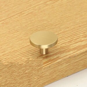 May include: A round, gold-toned cabinet knob with a smooth, flat surface and a cylindrical base.