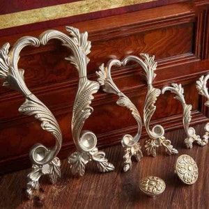 Könnte beinhalten: Fünf verzierte, silberfarbene Schranktürgriffe mit einem dekorativen Rollendesign. Die Griffe sind in einer Reihe vom größten zum kleinsten angeordnet. Zwei kleinere goldfarbene Knöpfe sind ebenfalls zu sehen.