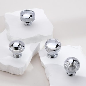 May include: Four chrome cabinet knobs with different designs. The knobs are displayed on white rock-like surfaces.
