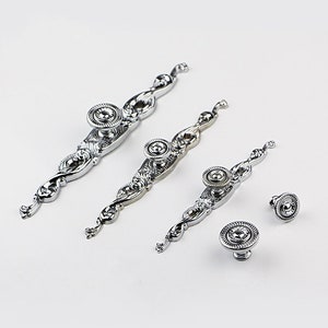 May include: Set of four silver-toned cabinet knobs with ornate designs. The knobs feature a round, raised center with a decorative pattern. The knobs are perfect for adding a touch of elegance to any home.