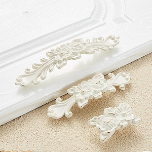 May include: Three white ornate drawer pulls with floral designs. The pulls are made of metal and have a distressed white finish.
