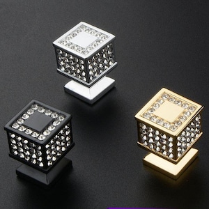 K&ouml;nnte beinhalten: Drei dekorative Schrankkn&ouml;pfe in Silber, Schwarz und Gold. Jeder Knopf hat ein quadratisches Design, das mit glitzernden Kristallen besetzt ist. Die Kn&ouml;pfe sind auf einer quadratischen Basis montiert und verleihen jedem M&ouml;belst&uuml;ck einen Hauch von Eleganz.