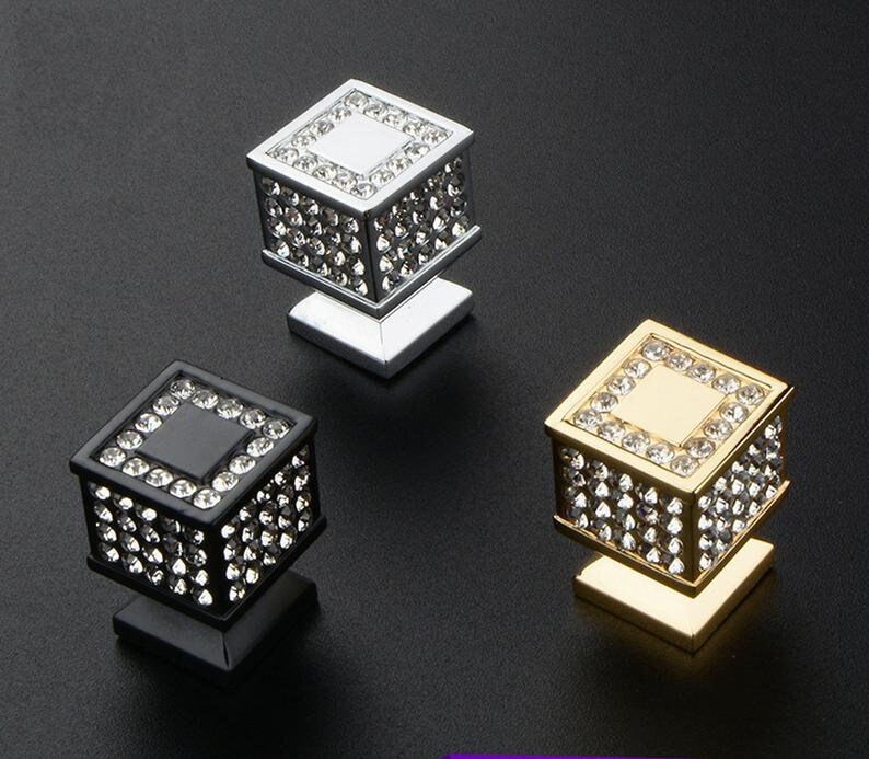 K&ouml;nnte beinhalten: Drei dekorative Schrankkn&ouml;pfe in Silber, Schwarz und Gold. Jeder Knopf hat ein quadratisches Design, das mit glitzernden Kristallen besetzt ist. Die Kn&ouml;pfe sind auf einer quadratischen Basis montiert und verleihen jedem M&ouml;belst&uuml;ck einen Hauch von Eleganz.