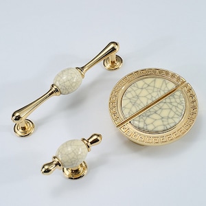 May include: Set of three gold cabinet knobs with a white crackle glaze finish. The knobs feature a round design with a Greek key border.