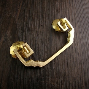 May include: Gold-toned metal drawer pull with a decorative, scalloped edge and a square design. The pull is designed to be mounted on a cabinet or drawer.