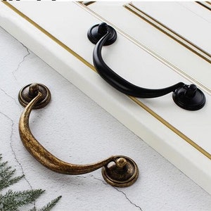 May include: Two cabinet door pulls, one black and one antique gold. The black pull has a simple, curved design. The antique gold pull has a more ornate, curved design.