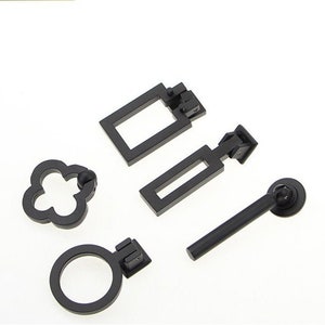 May include: Four black metal cabinet door pulls in different styles. The pulls include a round ring, a clover shape, a rectangular pull with a hinge, and a long bar pull with a round knob.