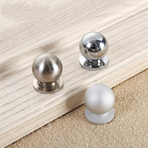 Op de afbeelding: Drie ronde kastknoppen in verschillende afwerkingen: zilver, geborsteld nikkel en chroom. De knoppen zijn allemaal even groot en hebben een glad, afgerond oppervlak.