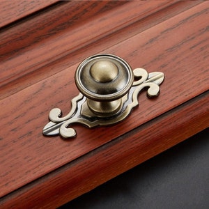 May include: A vintage-style cabinet knob with a round, brass-colored base and a decorative, ornate, scroll-shaped plate. The knob has a slightly aged, antique finish.