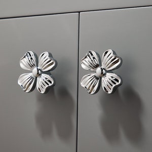 May include: Two silver flower-shaped cabinet knobs with a white centre. The knobs are attached to a grey cabinet door.