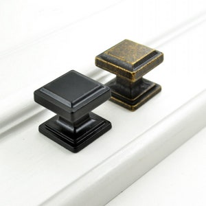 May include: Two square cabinet knobs, one black and one gold, with a slightly raised edge around the top and bottom. The knobs are made of metal and have a vintage look.