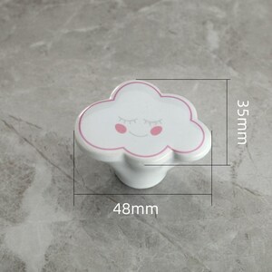 May include: White cloud-shaped cabinet knob with a pink outline and smiling face. The knob measures 48mm wide and 35mm high. A decorative accent for furniture, this knob adds a whimsical touch to drawers or cabinets.