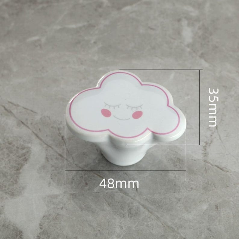 May include: White cloud-shaped cabinet knob with a pink outline and smiling face. The knob measures 48mm wide and 35mm high. A decorative accent for furniture, this knob adds a whimsical touch to drawers or cabinets.