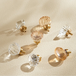 Puede incluir: Surtido de ocho pomos de armario de vidrio con bases de metal dorado. Los pomos son de varias formas y tamaños, incluyendo redondos, cuadrados y en forma de diamante. Algunos pomos son de vidrio transparente, mientras que otros son de color ámbar claro.