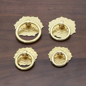 May include: Four gold-toned metal cabinet door pulls with a ring handle and a decorative floral design. The pulls are arranged in two rows of two.