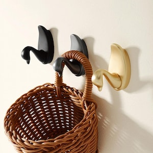 May include: Three decorative swan-shaped hooks mounted on a white wall. The hooks are black, gold, and silver. A brown wicker basket hangs from one of the hooks.