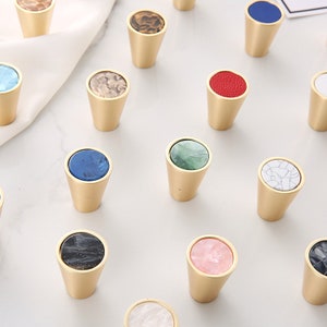 Puede incluir: Surtido de pomos decorativos para armarios con una variedad de incrustaciones coloridas y estampadas. Los pomos están hechos de latón y tienen una parte superior redonda y plana con un borde elevado. Las incrustaciones están hechas de diferentes materiales, incluyendo mármol, ágata y nácar.