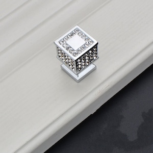 May include: A square cabinet knob with a chrome finish and a crystal-like design. The knob has a square base and a raised square top with a smaller square in the centre.