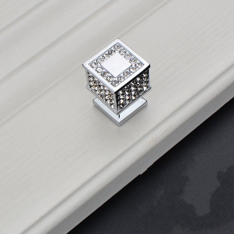 May include: A square cabinet knob with a chrome finish and a crystal-like design. The knob has a square base and a raised square top with a smaller square in the centre.
