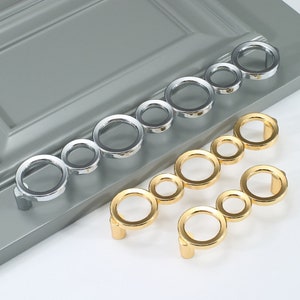 Könnte beinhalten: Zwei Schrankgriffe mit kreisförmigem Design. Ein Griff ist silberfarben und der andere goldfarben.
