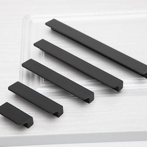 Può includere: Una collezione di maniglie per mobili nere opache di varie dimensioni. Le maniglie hanno un design rettangolare con un'area di presa incassata sul lato inferiore. L'hardware è disposto su una superficie bianca, mostrando le diverse lunghezze, misurate in centimetri.