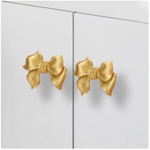 May include: Two gold bow-shaped cabinet knobs on a white cabinet door.