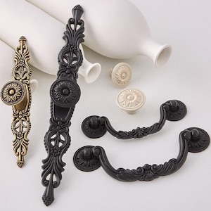 May include: A set of decorative cabinet hardware featuring ornate, floral designs. The set includes two black drawer pulls, two black cabinet pulls, and two knobs in antique gold and off-white.
