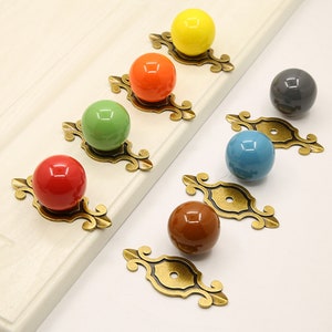 Könnte beinhalten: Satz von sieben Keramik-Schrankknöpfen in verschiedenen Farben, darunter Gelb, Orange, Grün, Rot, Blau, Braun und Grau. Jeder Knopf ist an einer goldfarbenen Metallbasis mit einem dekorativen Design befestigt.