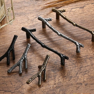 Pode incluir: Cinco estilos diferentes de botões de armário em preto, prata e bronze com forma de galhos. Os botões são de metal e têm um aspeto rústico e natural.