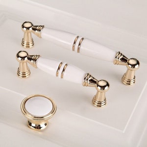 May include: Three white ceramic cabinet knobs with gold accents. Two knobs are long and rectangular with a gold trim around the edges. The third knob is round with a gold base.