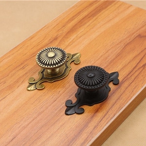 May include: Two decorative cabinet knobs with ornate metal designs. One knob is gold with a sunburst pattern, the other is black with a similar pattern.