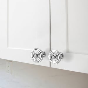 May include: Two silver round cabinet knobs with a concentric circle design. The knobs are mounted on white cabinet doors.