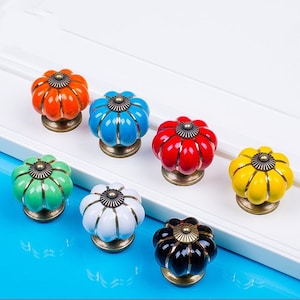 Könnte beinhalten: Sortiment von sieben Keramik-Schrankknöpfen in Form von Kürbissen in verschiedenen Farben, darunter Orange, Blau, Rot, Grün, Weiß, Gelb und Schwarz. Jeder Knopf hat eine goldfarbene Metallbasis.