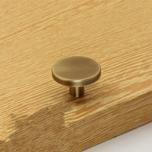 May include: A round, antique brass cabinet knob with a smooth, slightly raised surface. The knob is mounted on a wooden surface.