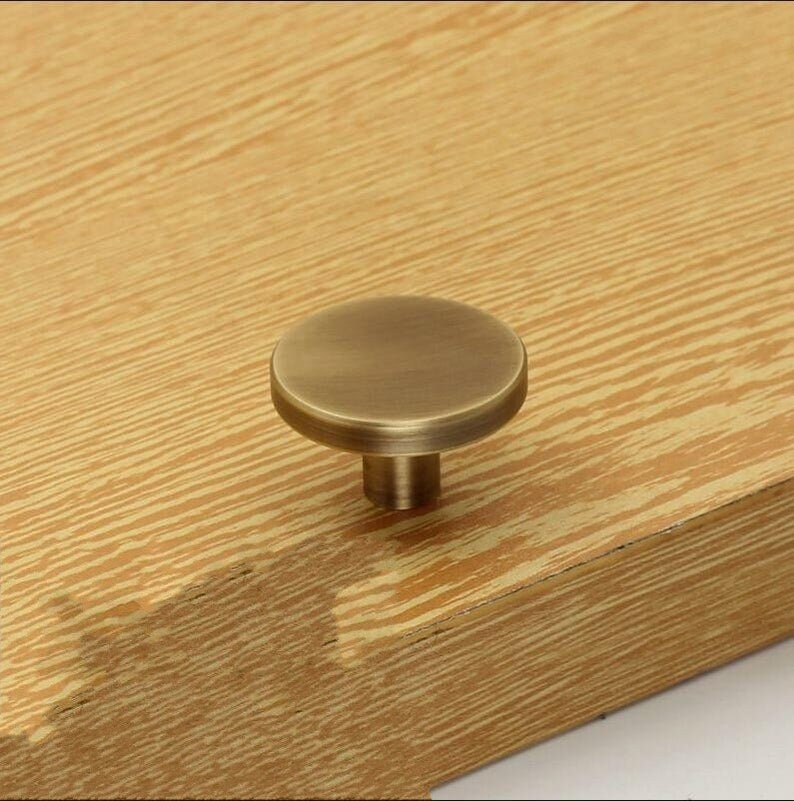 May include: A round, antique brass cabinet knob with a smooth, slightly raised surface. The knob is mounted on a wooden surface.