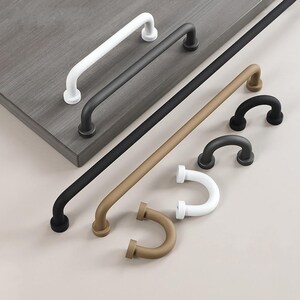 Peut inclure: Une sélection de poignées de meubles de différentes couleurs et styles. Les poignées sont blanches, noires et beiges, dans des modèles droits et courbes. Les poignées sont en métal et sont présentées sur une surface grise.