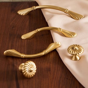 Puede incluir: Cuatro tiradores de armario de color dorado con un diseño decorativo acanalado. Dos son tiradores arqueados y dos son pomos redondos.