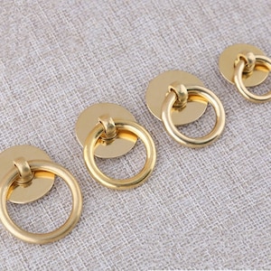 May include: Four gold-toned metal ring pulls of varying sizes. The ring pulls have a smooth, polished finish and are designed for use on cabinets, drawers, or doors.