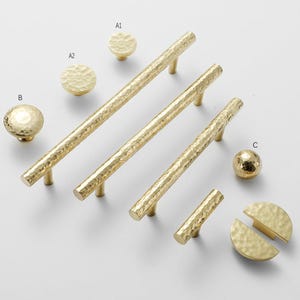 Könnte beinhalten: Eine Sammlung goldfarbener Schrankbeschläge, darunter Knöpfe und Griffe. Die Beschläge haben eine gehämmerten Textur und sind auf einer weißen Oberfläche angeordnet. Die Kollektion umfasst verschiedene Größen und Formen.