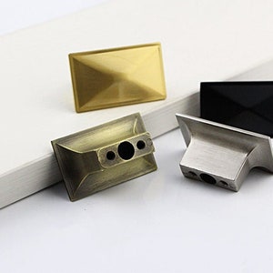 Puede incluir: Tres pomos de armario en diferentes acabados: dorado, lat&oacute;n antiguo y n&iacute;quel cepillado. Cada pomo tiene una forma rectangular con un centro ligeramente elevado.