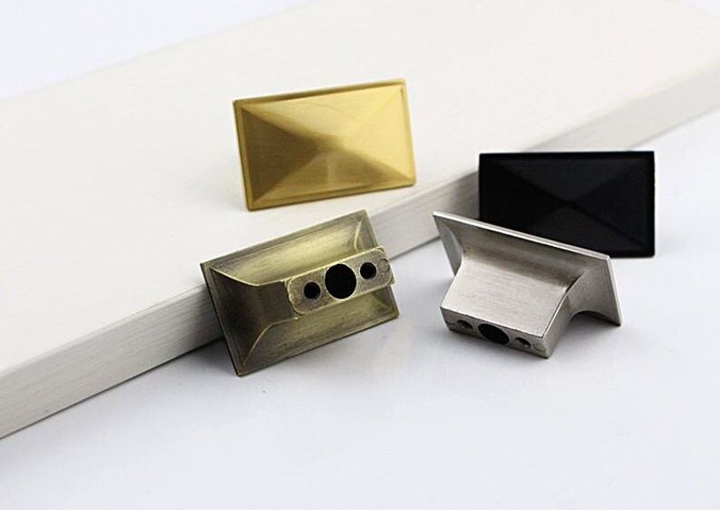 Puede incluir: Tres pomos de armario en diferentes acabados: dorado, lat&oacute;n antiguo y n&iacute;quel cepillado. Cada pomo tiene una forma rectangular con un centro ligeramente elevado.