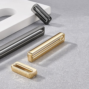 May include: Four cabinet pulls in different finishes: black, grey, gold, and gold. The pulls are rectangular with a cutout in the centre.