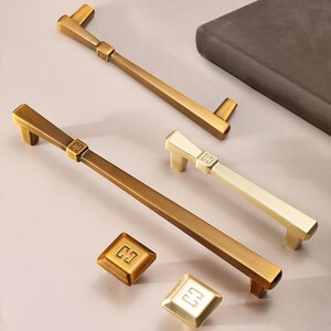 May include: Five cabinet hardware pulls in gold and brass finishes. The pulls are all rectangular with a square knob at the end. The pulls have a decorative design with the letters "GG" engraved on the knob.