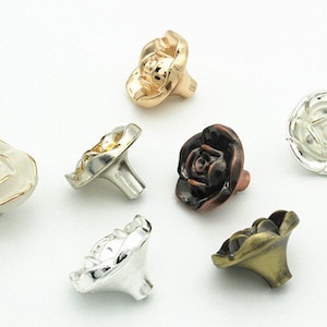 Op de afbeelding: Assortiment van zes roosvormige kastknoppen in verschillende afwerkingen, waaronder goud, zilver, wit en brons.