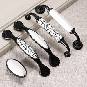 Pode incluir: Quatro botões de armário pretos com frentes de cerâmica branca com um padrão geométrico. Os botões têm uma forma retangular arredondada com uma ligeira curva na parte superior.