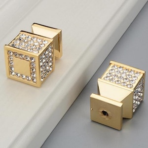 K&ouml;nnte beinhalten: Zwei goldfarbene Schrankkn&ouml;pfe mit quadratischem, w&uuml;rfelf&ouml;rmigem Design. Die Kn&ouml;pfe sind mit kleinen, klaren Kristallen besetzt und haben eine quadratische Aussparung auf der Vorderseite. Die Kn&ouml;pfe sind auf einer wei&szlig;en Oberfl&auml;che montiert.