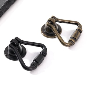 以下が含まれることがあります： リング型のハンドルと装飾的なベースが付いた、黒とアンティークブラスの2つのキャビネットドアプルです。